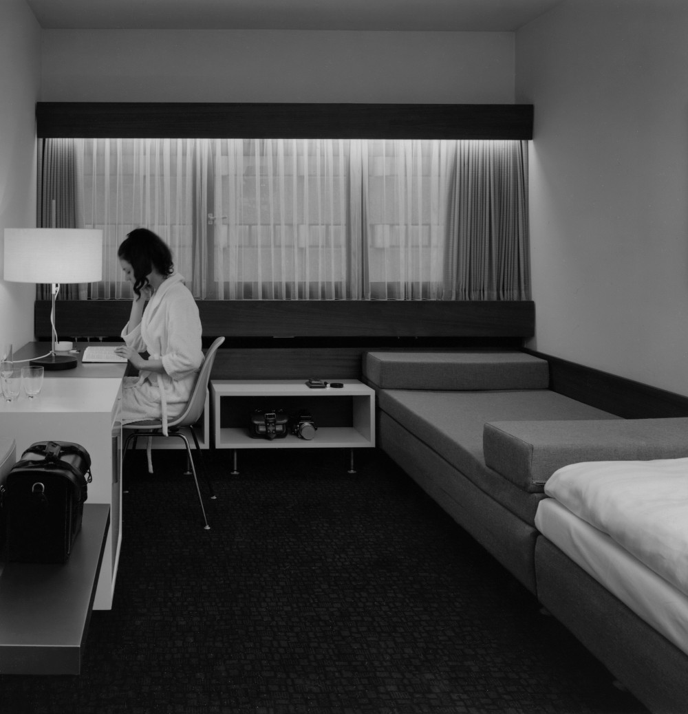 Schwarz-Weiß Fotografie: Blick in ein Hotelzimmer mit einer Person in weißem Bademantel, die an einem Tisch sitzt und liest